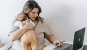 A woman sits holding a baby to her chest and looking at a laptop.