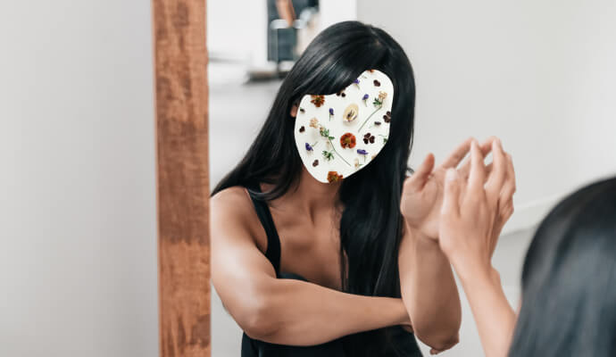 A woman with a floral collage over her face sits looking at her reflection in the mirror.